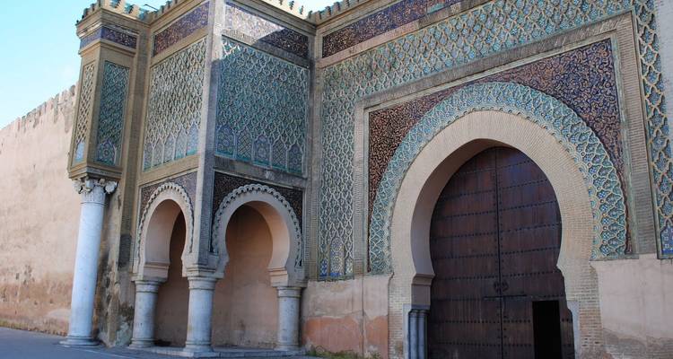 Une porte de ville marocaine ornée avec un travail de carrelage complexe, de grandes portes en bois cintrées et des colonnes classiques sous un ciel bleu dégagé.