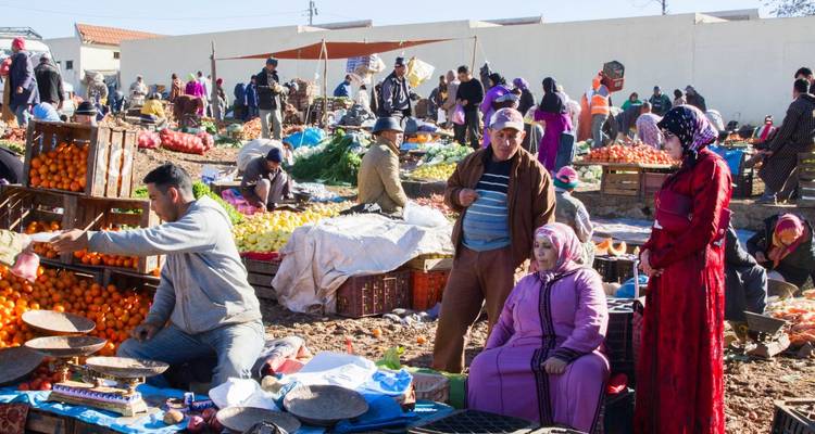 Une scène animée de marché extérieur au Maroc avec des vendeurs proposant des produits colorés, des clients qui parcourent les étals et des interactions dynamiques partout.