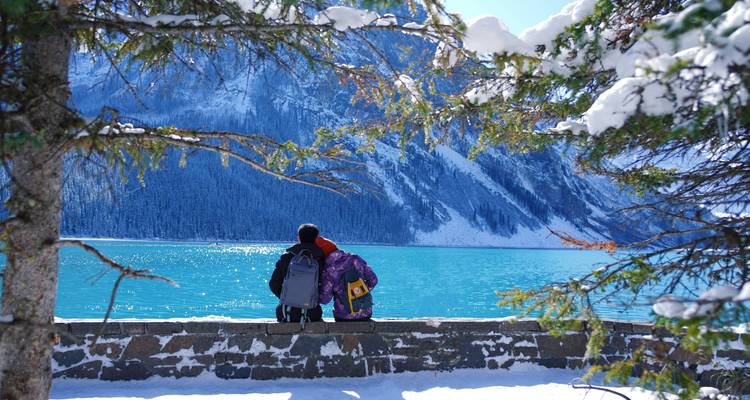 Un couple avec des sacs à dos est assis sur un banc en pierre, admirant les eaux turquoise du lac Louise encadrées par des montagnes enneigées.