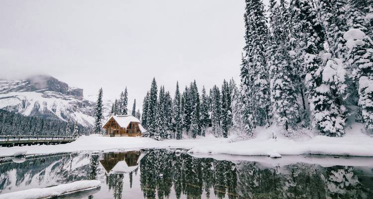 Les sapins persistants chargés de neige se reflètent dans les eaux cristallines à côté d'une cabane en bois rustique sur le lac Émeraude.