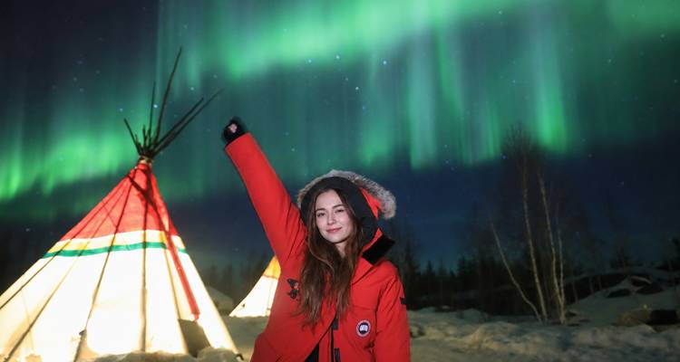Femme souriante en parka rouge posant le bras levé sous les aurores boréales d'un vert éclatant à côté d'un tipi illuminé sur un sol enneigé.
