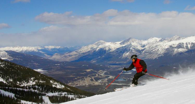 Skieur en veste rouge dévale une pente enneigée avec de vastes chaînes de montagnes Rocheuses et une vallée en contrebas.