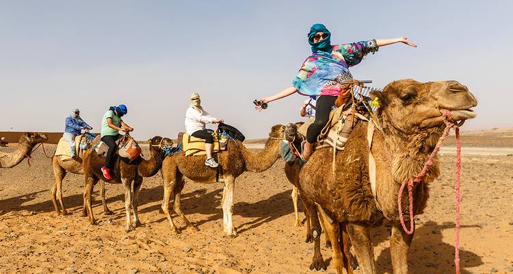 Caravane de touristes chevauchant des chameaux à travers les sables dorés du désert, un cavalier posant les bras écartés.