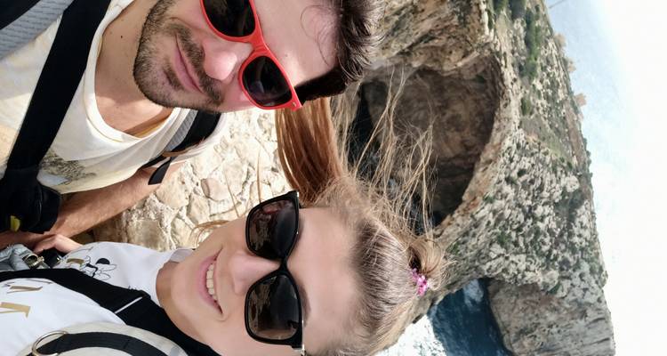 Selfie van een glimlachend stel met zonnebrillen met de Blue Grotto zeenatuurlijke boog achter hen.