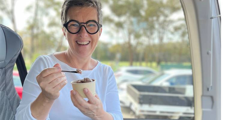 Une femme portant des lunettes savoure une tasse de gelato au chocolat à l'intérieur d'un bus touristique avec des voitures visibles à l'extérieur.