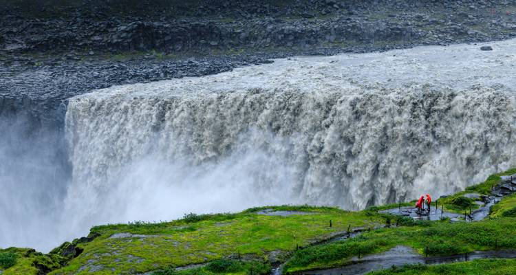 Immense mur d'eau de Dettifoss avec deux visiteurs vêtus de vestes imperméables pour donner l'échelle.