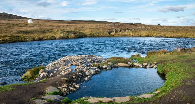 Petite piscine thermale au bord de la rivière avec un paysage vallonné et un vaste ciel en arrière-plan.