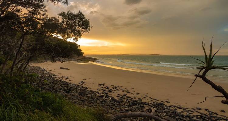 Playa rocosa y tranquila al atardecer con luz dorada cálida reflejándose en el agua tranquila y las nubes.