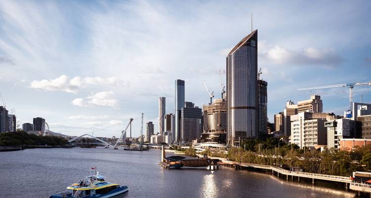 El moderno skyline de Brisbane a lo largo del río con un ferry y puentes peatonales bajo un cielo brillante.