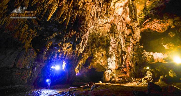Stalactites illuminées dans une grande chambre de grotte avec un visiteur solitaire, filigrane de marque dans le coin.