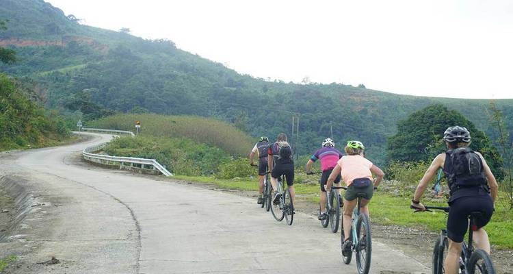 Les cyclistes montent une route rurale sinueuse en béton à travers des collines verdoyantes sous une lumière nuageuse.