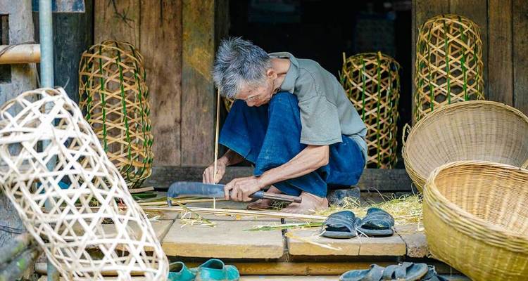 Un artisan âgé accroupi tisse des bandes de bambou en paniers sur un porche en bois.