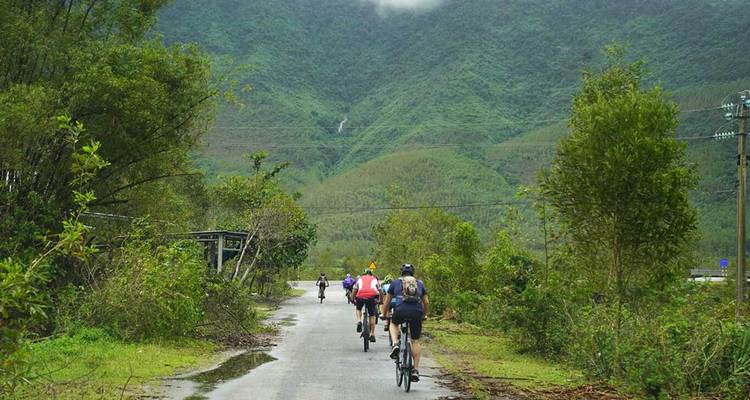 Les cyclistes pédalent le long d'une route rurale mouillée bordée par une végétation luxuriante et des collines lointaines après la pluie.