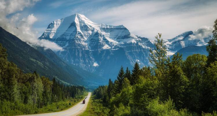 Der schneebedeckte Mount Robson erhebt sich über einer bewaldeten Straße, die sich in die Ferne erstreckt.