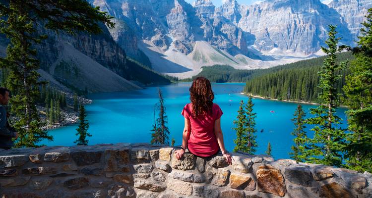 Der Reisende sitzt auf einer Steinmauer und blickt über den leuchtend blauen Moraine Lake und die rauen Gipfel.