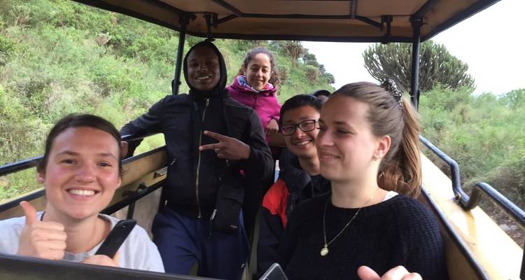 Een groep glimlachende reizigers zit in een open safariauto en steekt duimen en vredestekens omhoog.