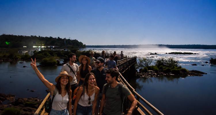Un groupe heureux marche sur une passerelle en bois aux chutes d'Iguazu sous un ciel éclairé par la lune avec du brouillard et de l'eau en cascade derrière.