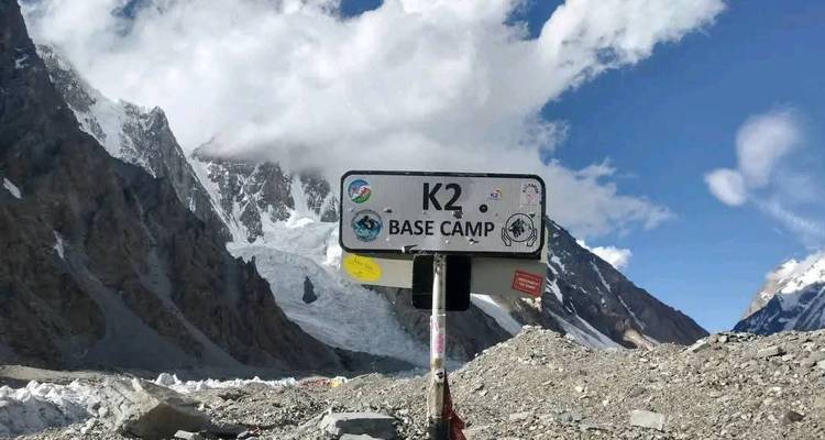 Panneau indicateur indiquant « K2 Base Camp » situé parmi un terrain glaciaire rocheux avec des montagnes spectaculaires couvertes de neige dominant en arrière-plan sous un ciel bleu éclatant.