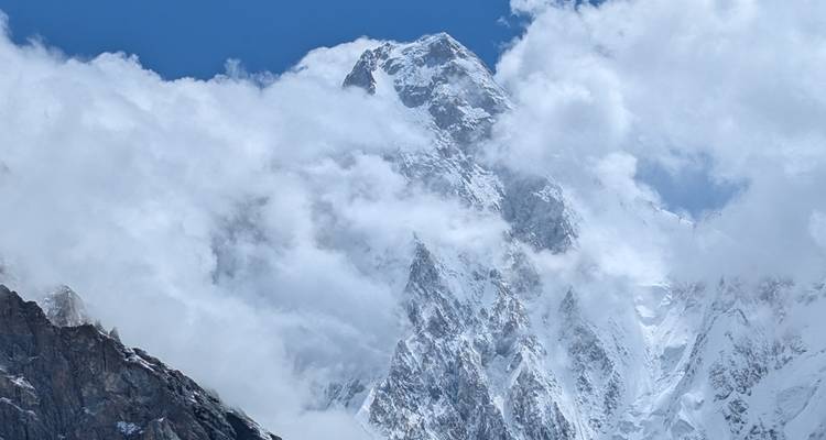 Sommet enneigé perçant les nuages tourbillonnants, révélant des parois glacées abruptes sur un ciel bleu profond.