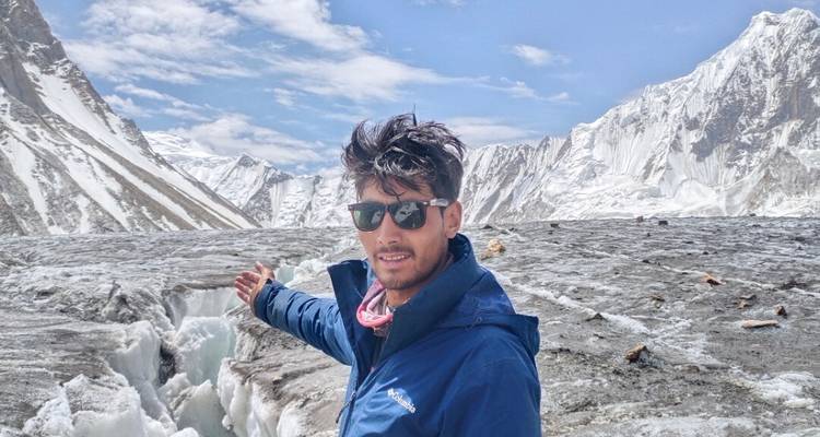 Jeune trekkeuse portant des lunettes de soleil faisant un geste en direction d'une crevasse sur un vaste glacier encadré par les pics enneigés du Karakoram.