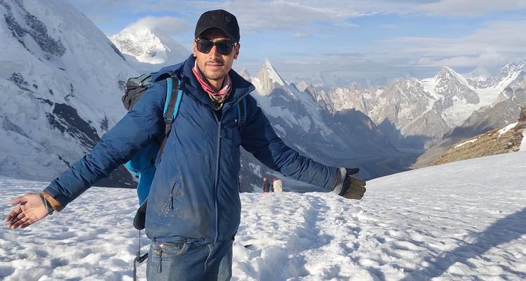 Trekkeur en veste bleue posant sur un col enneigé avec les pics dramatiques du Karakoram se dressant majestueusement à l'arrière-plan.