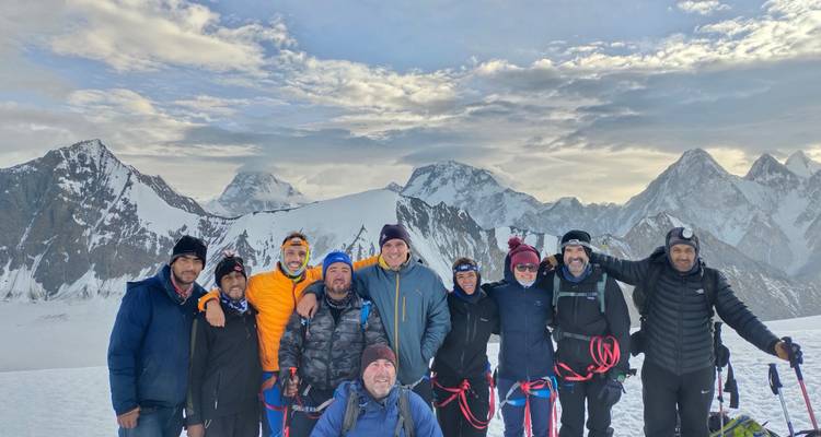 Équipe d'expédition souriante en tenue d'escalade posant ensemble sur un glacier avec des pics imposants en arrière-plan.