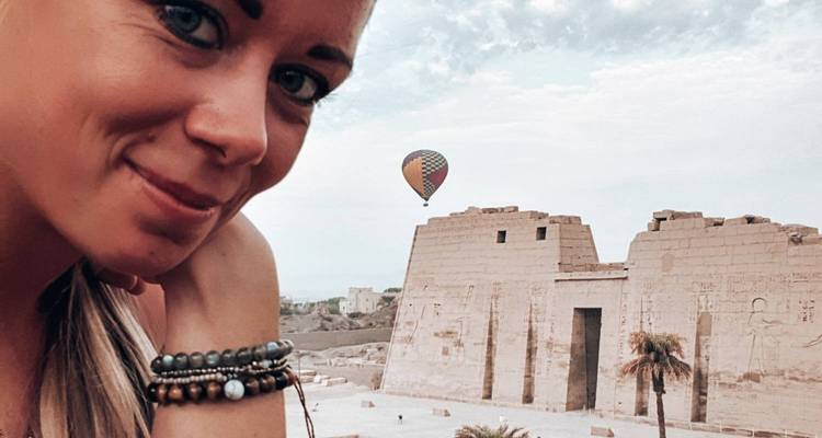 Close-up selfie of a woman with ancient temple ruins and a distant hot-air balloon behind her.