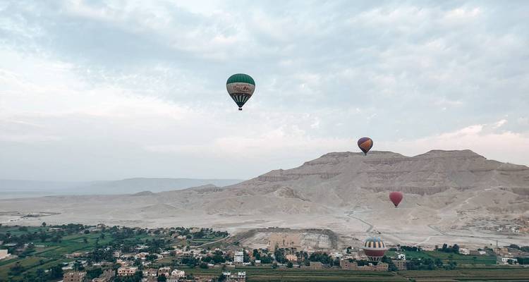 Several balloons hover over farmland and desert foothills beneath a pale morning sky.