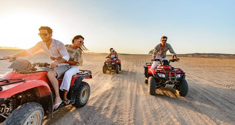 Grupo de jóvenes viajeros conduciendo motos cuadriclos rojas a través de las arenas del desierto al atardecer