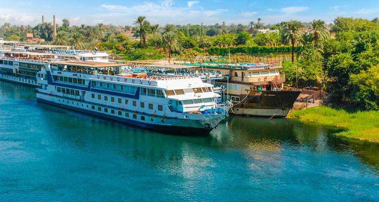 Múltiples cruceros del Nilo atracados uno al lado del otro en el río azul con una orilla bordeada de palmeras