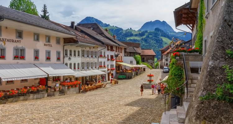 Encantadora calle principal adoquinada de Gruyères llena de restaurantes decorados con flores y picos alpinos de fondo
