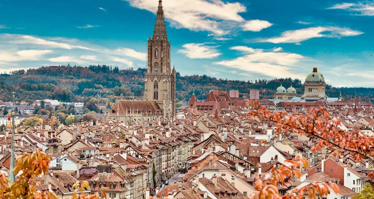 Vista panorámica del casco antiguo de Berna con la alta aguja de la catedral y el Palacio Federal con cúpula rodeado de tejados de tejas y colinas boscosas bajo un cielo azul brillante.