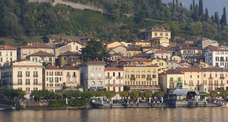 Fila de edificios y hoteles de colores pastel a orillas del lago reflejados en las aguas tranquilas del Lago Como con colinas boscosas elevándose detrás.