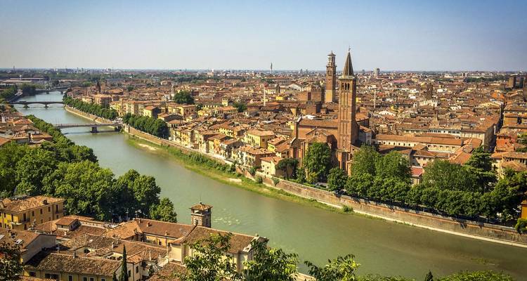 Vista elevada de los tejados de terracota de Verona bordeando el río Adigio con puentes arqueados y altos campanarios de iglesias bajo un cielo despejado.