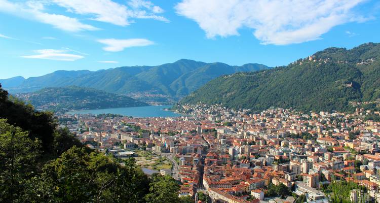 Panorama espectacular desde arriba de Como mostrando bloques de ciudad con techos rojos, el Lago Como y las montañas verdes que lo rodean bajo un cielo azul brillante.