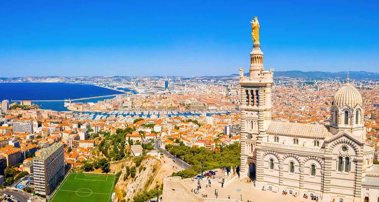 Vista aérea panorámica de la ciudad de Marsella, el Viejo Puerto y la basílica de Notre-Dame de la Garde bajo un cielo azul radiante.