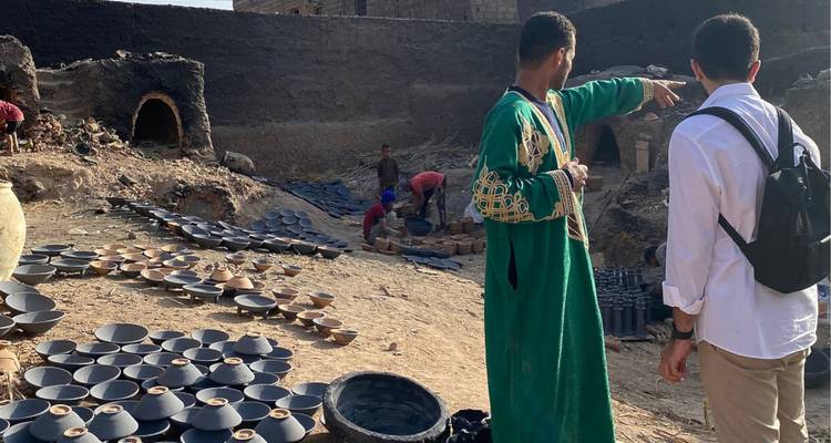 Un guía con una túnica verde le muestra a un visitante hornos de cerámica tradicional y filas de cuencos de arcilla secándose al sol.
