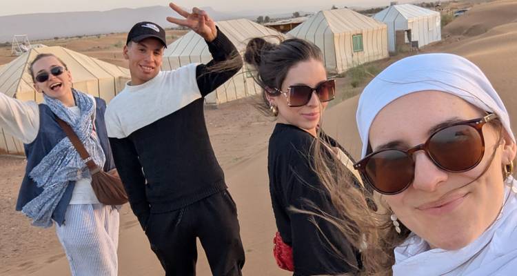 Cuatro amigos se toman una selfie en una duna ventosa cerca de un campamento desértico de lona.