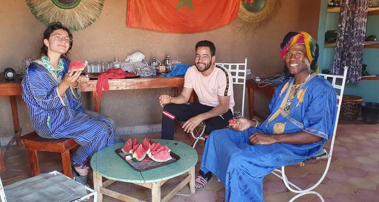Los viajeros disfrutan de una sandía fresca con un anfitrión local dentro de una habitación de adobe decorada con textiles marroquíes.