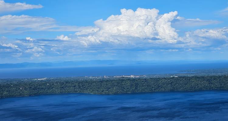 Expansive aerial view over deep blue lake, dense green jungle shore and distant mountains beneath billowing white clouds.