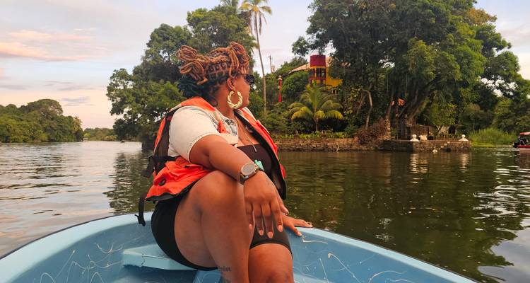 Woman in life jacket relaxes on a small blue boat cruising calm jungle waterways past tree-covered islets and colorful houses.