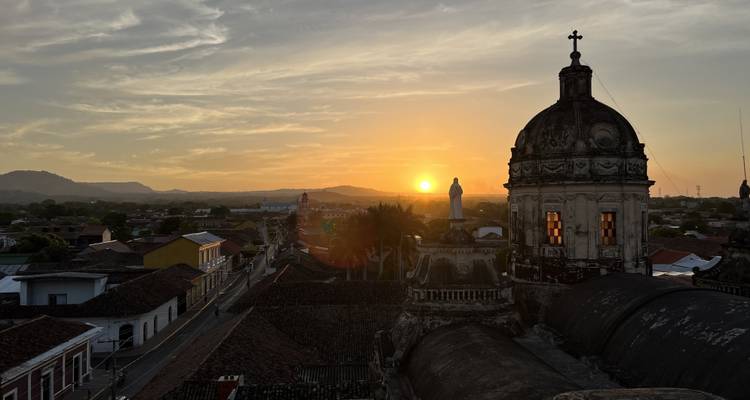 Sunset over Granada with silhouetted church dome and statue glowing windows atop tiled rooftops against orange sky and distant hills.