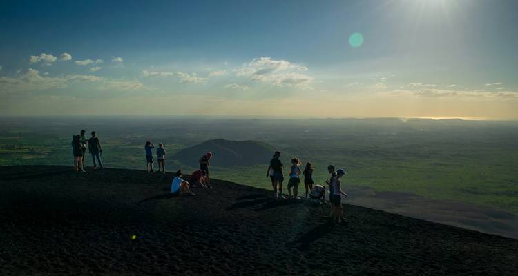 Small groups of hikers stand atop a dark volcanic cone enjoying vast green plains and shining ocean in the distance.