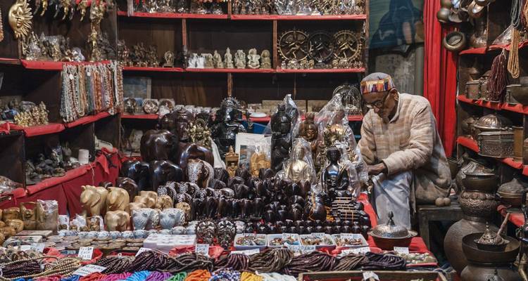 Un vendedor mayor organiza figurillas talladas y joyas en un puesto de mercado abarrotado lleno de artesanías coloridas.
