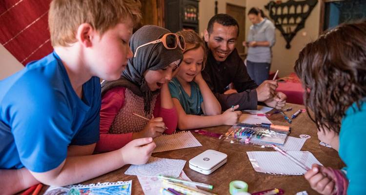 Niños y un guía local escribiendo y dibujando juntos con entusiasmo alrededor de una mesa baja en un riad.