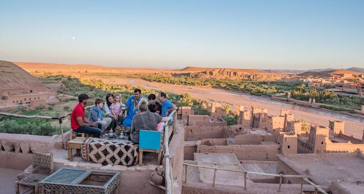 Un grupo familiar disfrutando del té en una terraza de azotea con vistas a las kasbahs de adobe y al valle verde de Ait Benhaddou al atardecer.
