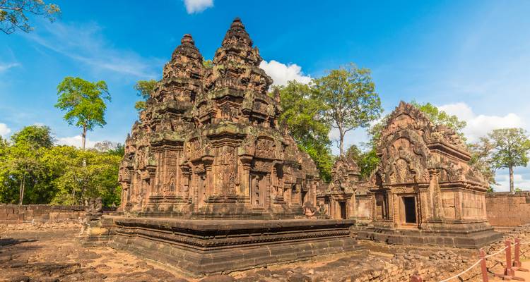 Des tours de temple khmer finement sculptées en pierre rose, entourées par une forêt verdoyante sous un ciel bleu dégagé.