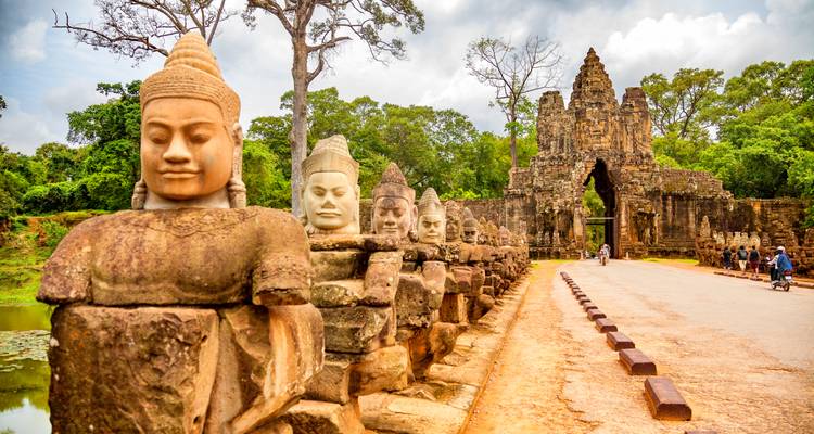 Des statues de gardiens en pierre bordent la route d'accès menant vers une ancienne porte d'Angkor avec des visiteurs marchant à proximité.