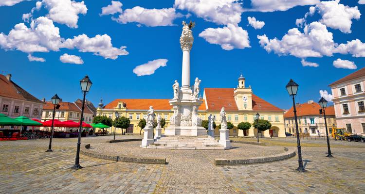 Plaza abierta con una columna de peste barroca blanca contra un cielo azul intenso y nubes.