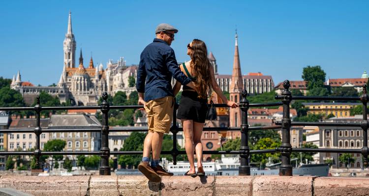 Un jeune couple se tient près d'une balustrade avec vue sur le Bastion des Pêcheurs de Budapest et l'horizon historique lors d'une journée ensoleillée.